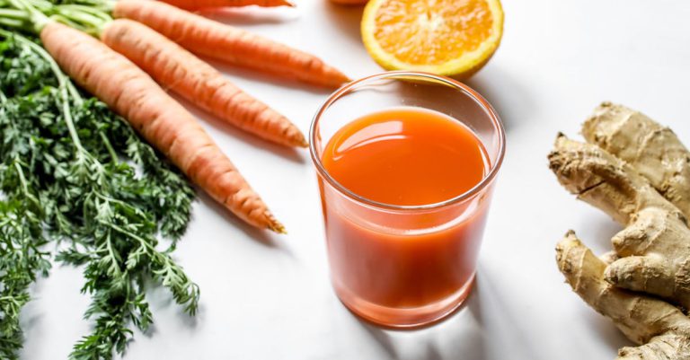 Fresh carrot ginger and orange juice surrounded by ingredients on a white background.