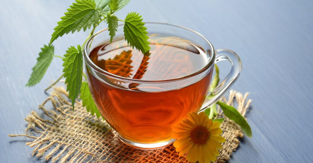 A refreshing glass cup of herbal tea garnished with nettle leaves and marigold flower.